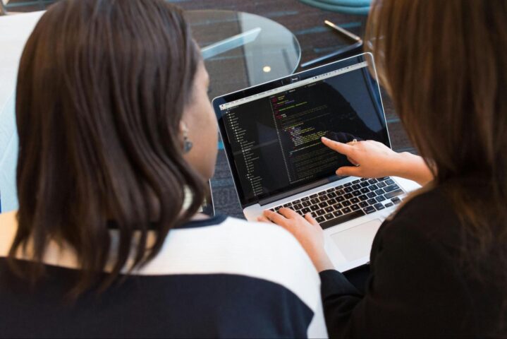 Two people collaborating on coding while looking at a laptop screen, with one person pointing at the code.