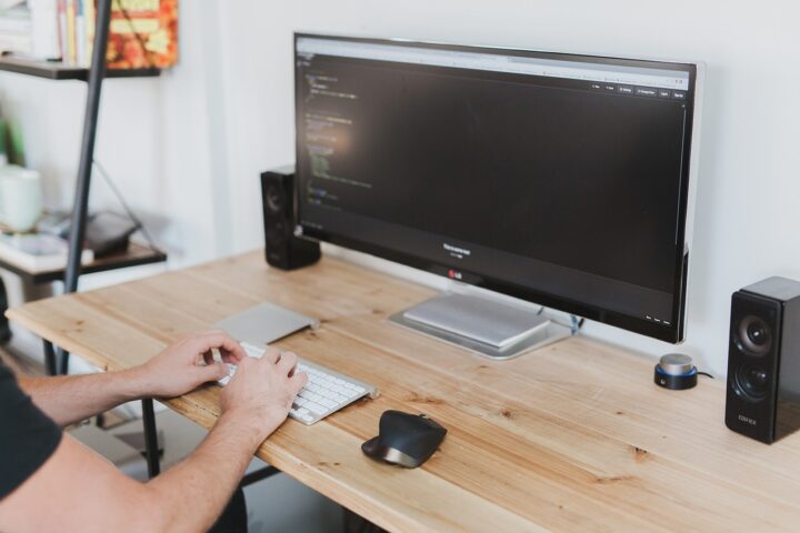 A developer sitting at a desk, typing on a keyboard with code displayed on a large monitor. The workspace is minimal and organized, focusing on coding.