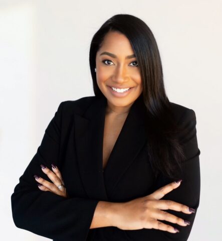 A woman with long black hair, wearing a black blazer, smiles confidently with arms crossed against a plain light background, embodying the professionalism and assurance associated with AI security best practices.